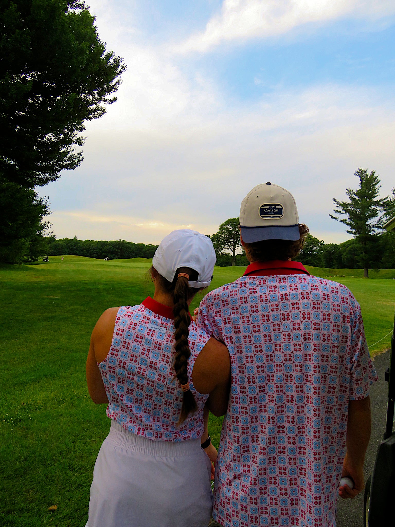 Two people standing on a golf course, seen from behind, wearing matching patterned shirts and white bottoms. The person on the right wears a beige baseball cap, and the background shows a green fairway under a blue sky with clouds.